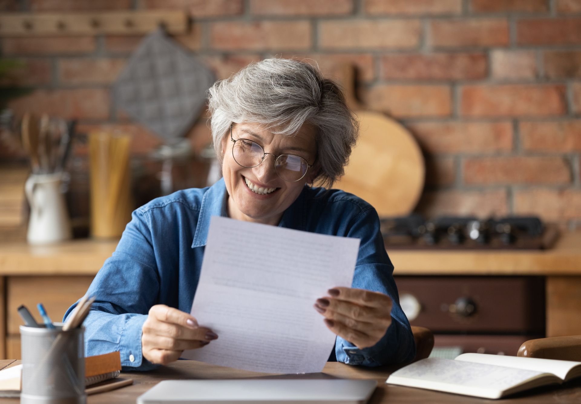 Smiling person in a blue shirt reading a sheet of paper at a kitchen table with an open book nearby