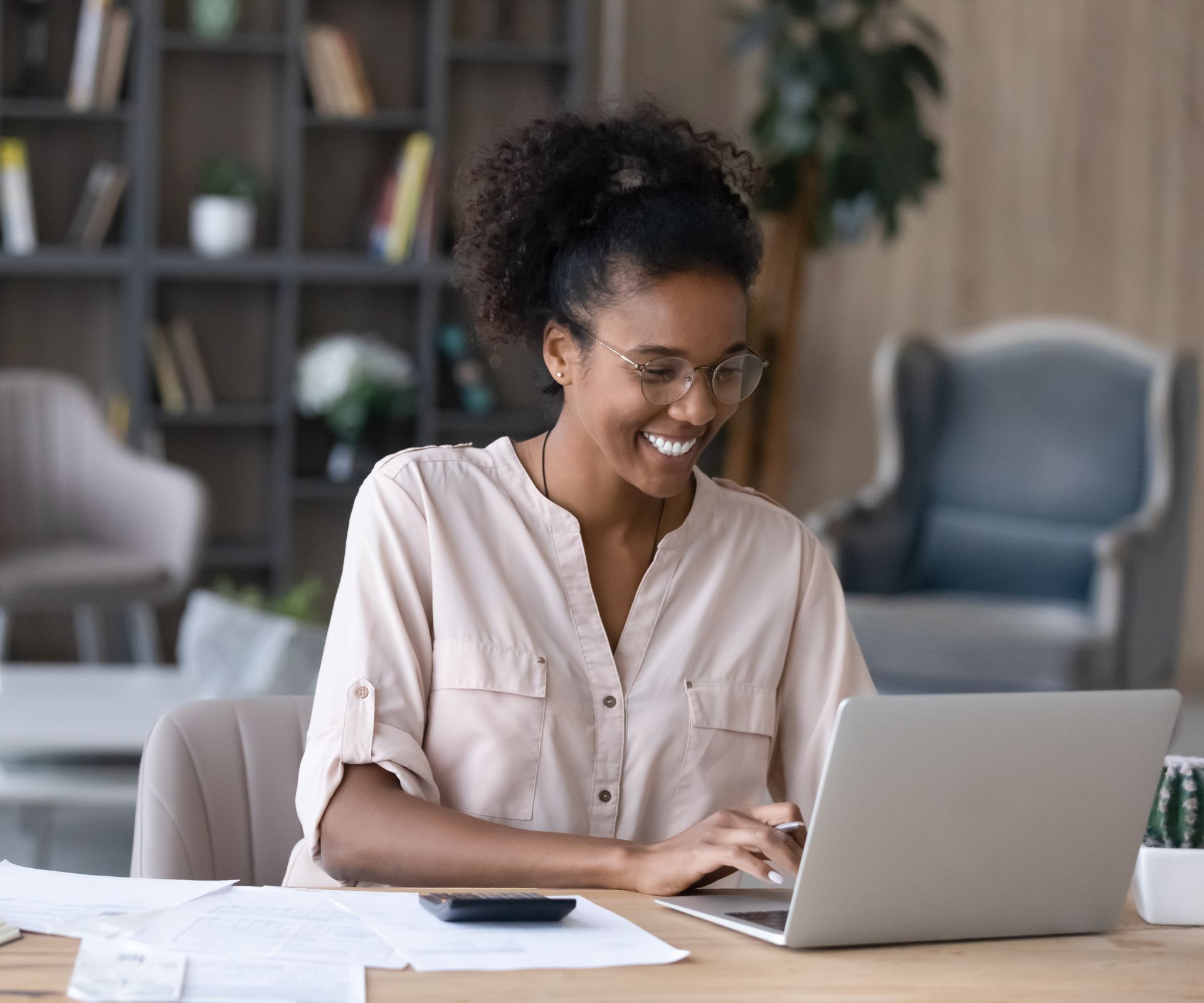 Woman smiling while typing on a laptop at a table in a bright living room.