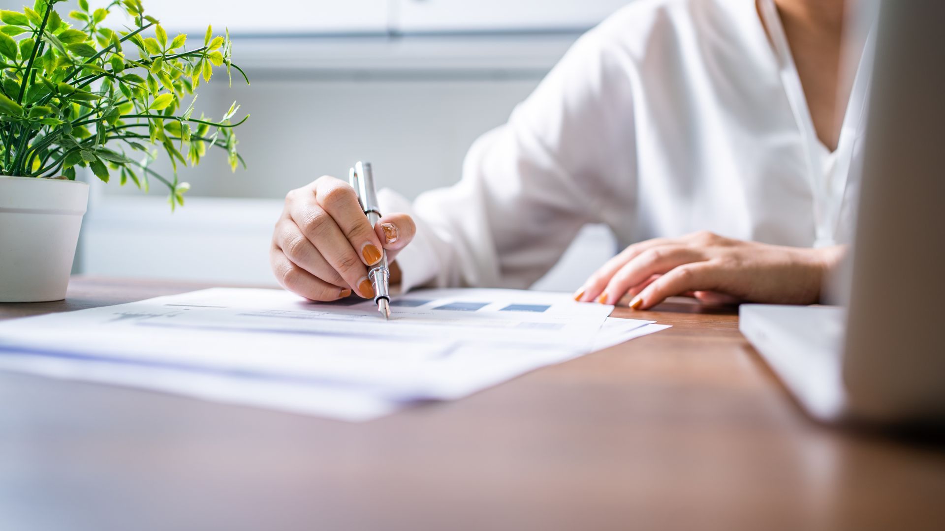 Person writing on papers at a desk beside a laptop and small potted plant.