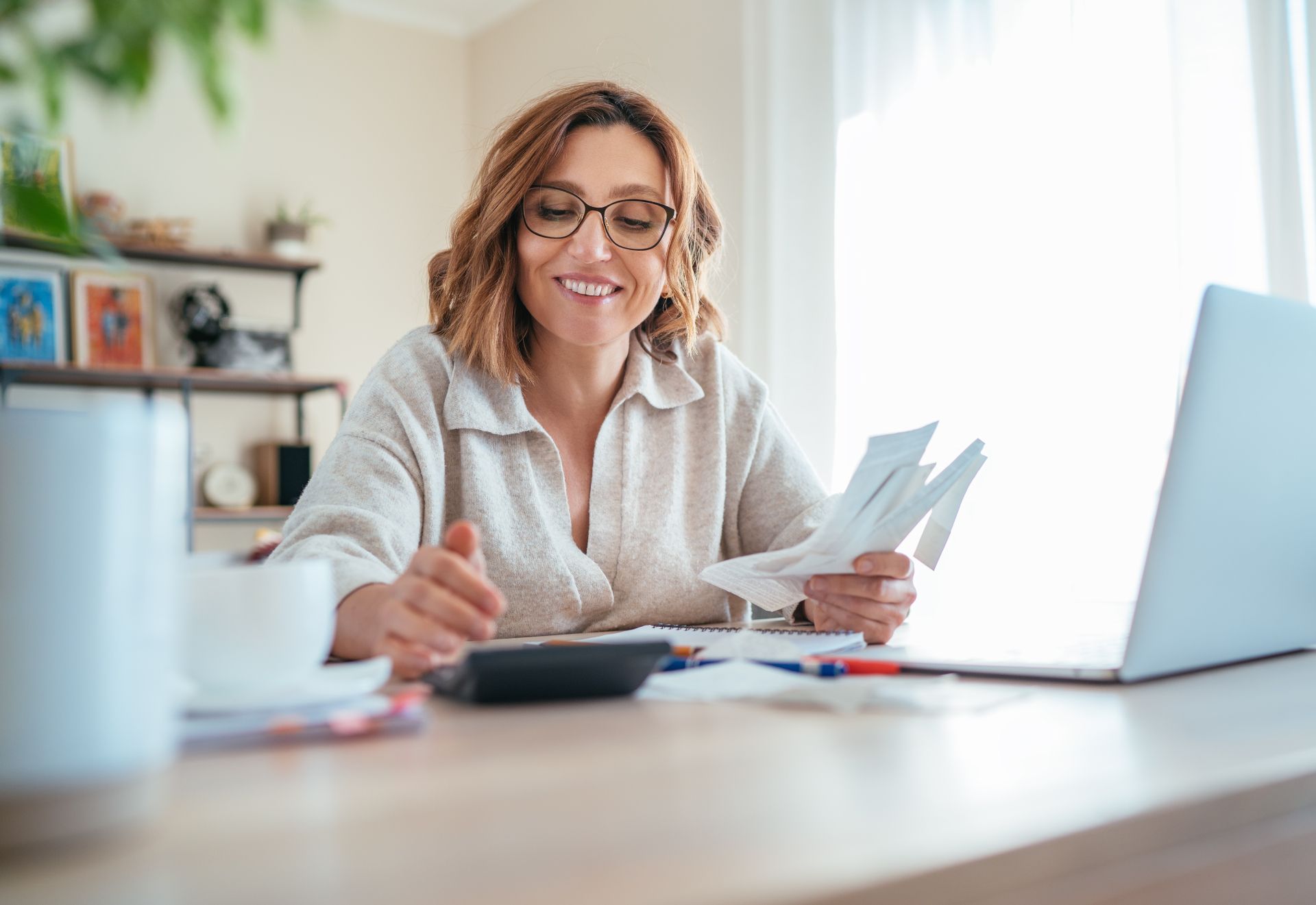 Woman smiling at a desk, working with papers beside a laptop and calculator in a bright office