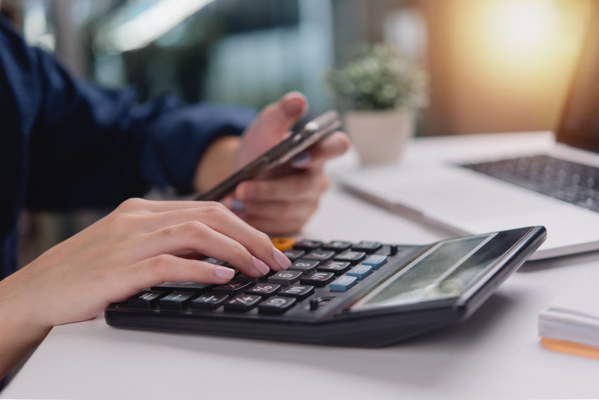 Hands using a calculator and smartphone at a desk with a laptop in the background