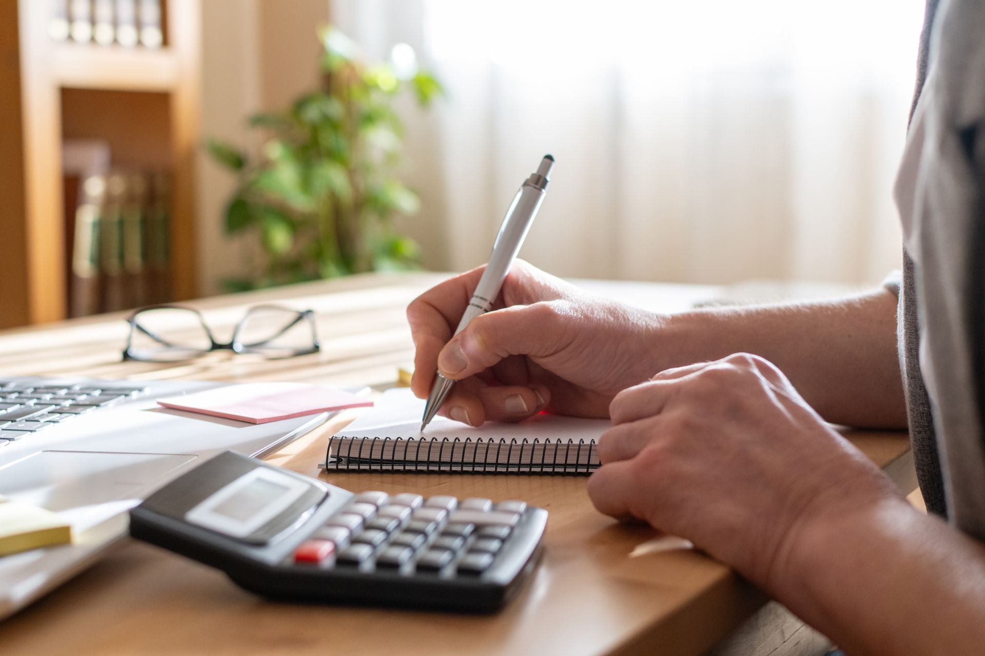 Hands writing in a notebook beside a calculator and laptop on a desk
