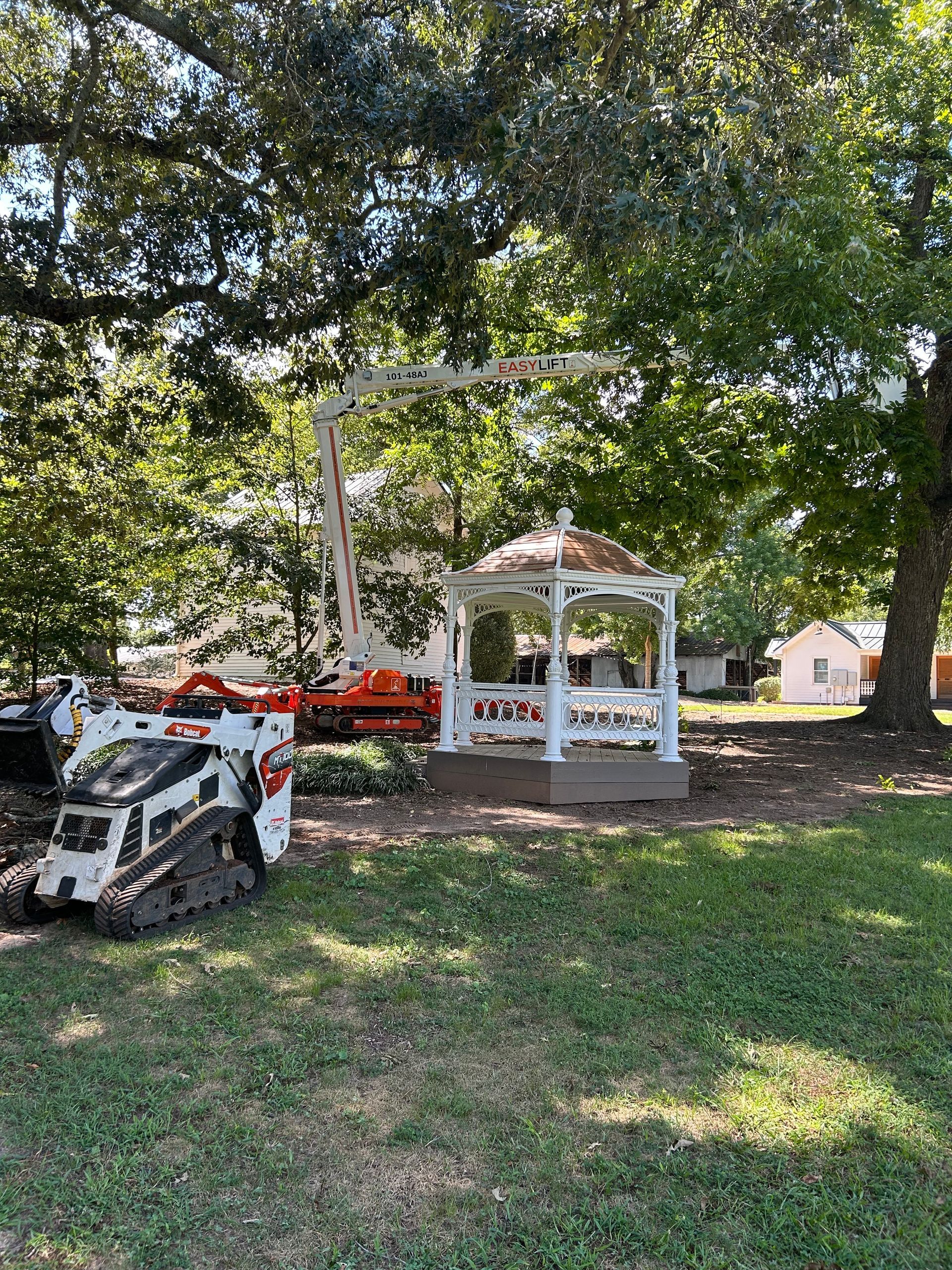 White gazebo next to a tracked skid steer and an orange lift under a large tree.