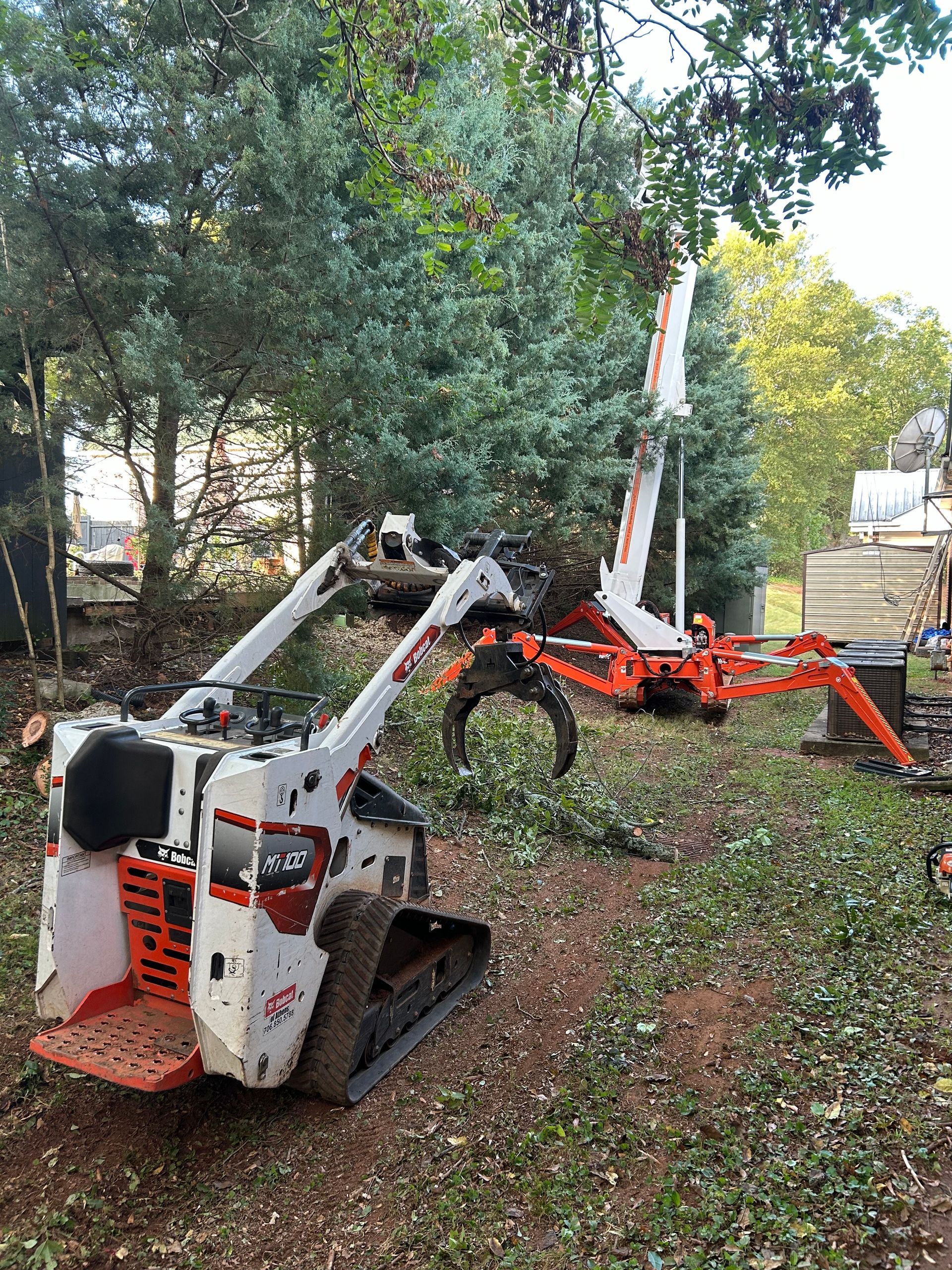 Two orange and white tree trimming machines working on a tree in a wooded area.