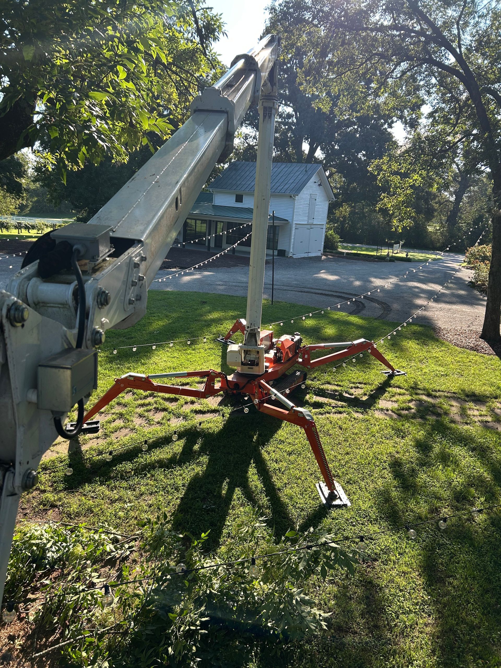 Orange spider lift on grass, arm extended toward white building in a park.