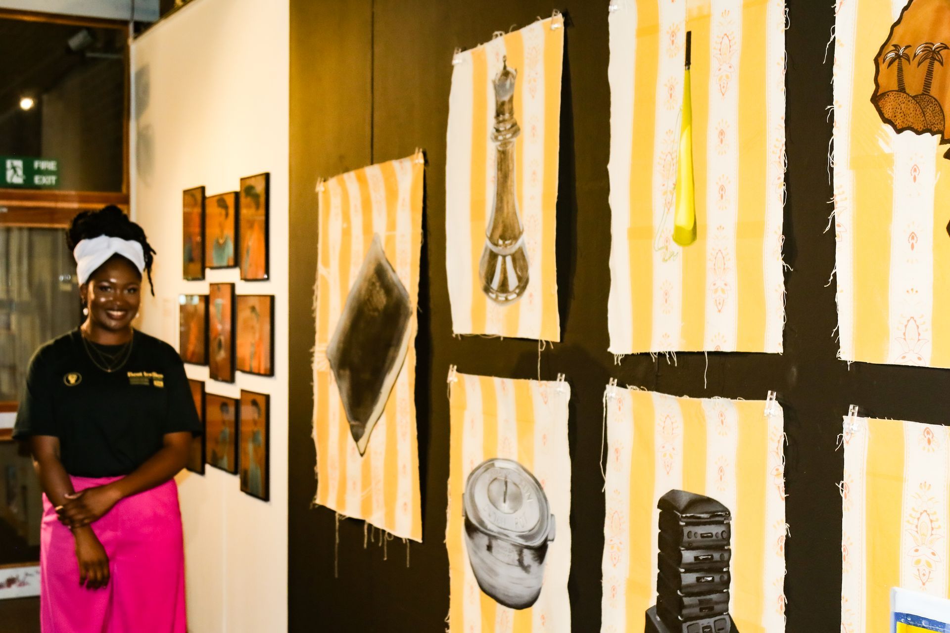 Woman in pink skirt stands near artwork in a gallery. Art is on walls; yellow & white stripes.
