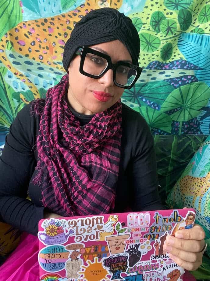 Woman wearing a black turban, glasses, and pink scarf, holding a laptop with stickers.