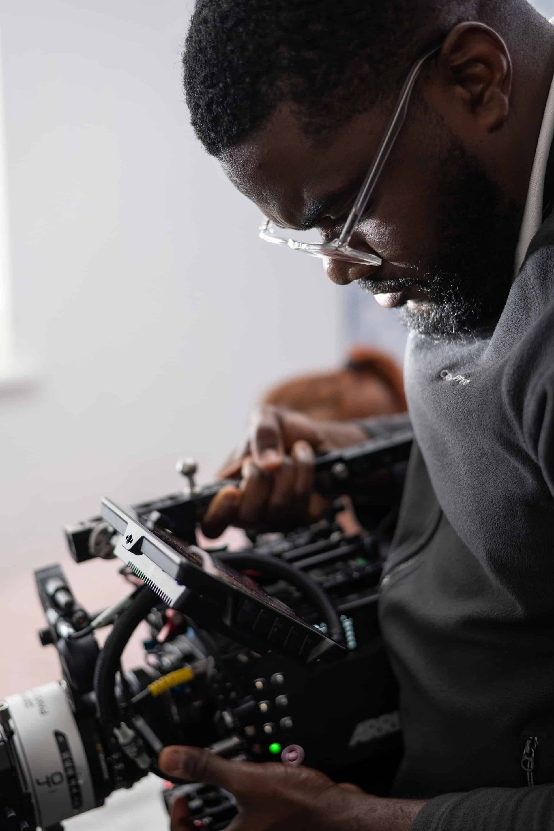 Man in glasses adjusts a video camera. He's indoors, likely filming.