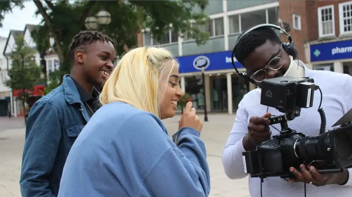 Filming scene: Three people laughing on a city street. A man operates a camera, focusing on two other people.