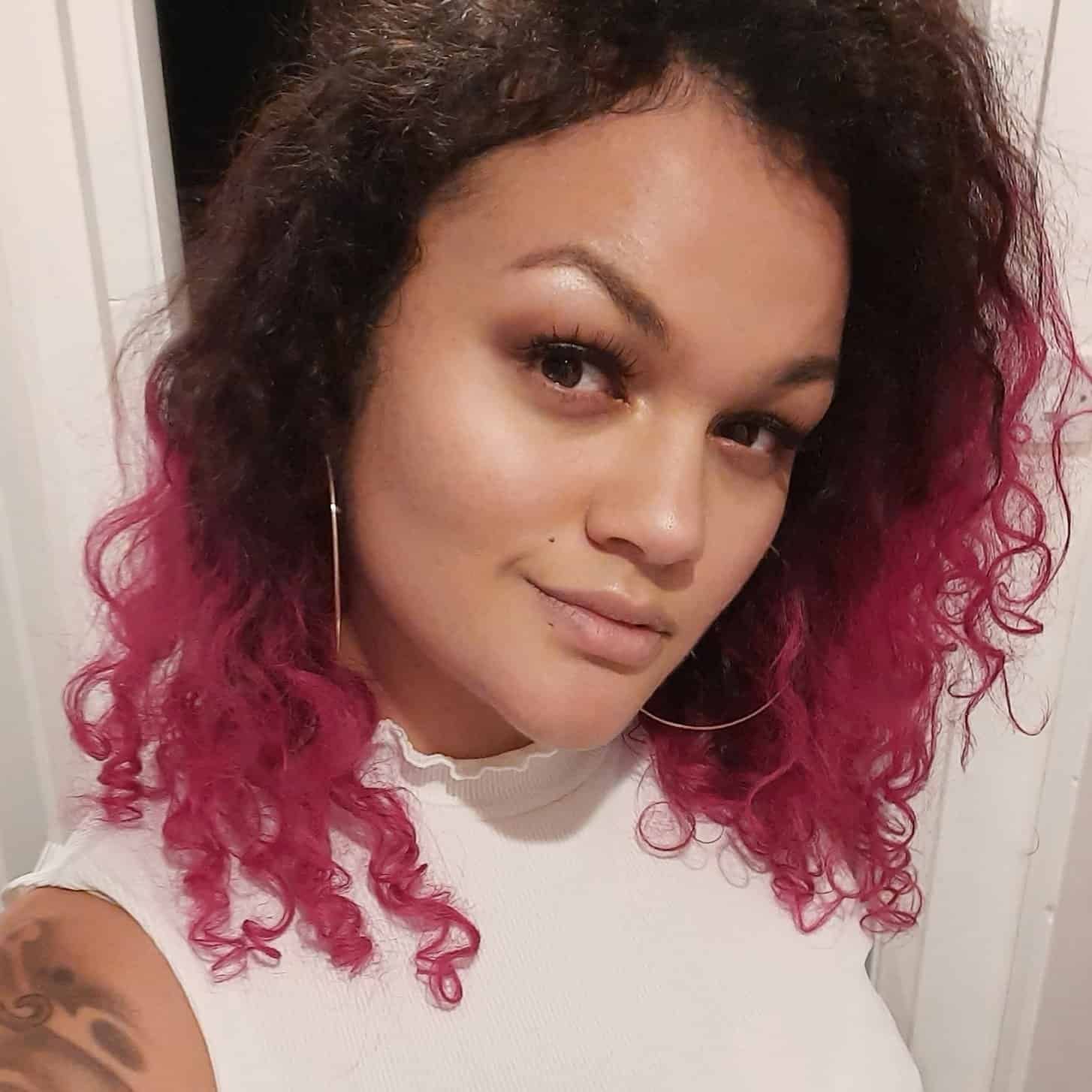 Woman with pink-tipped curly hair, wearing a white top and hoop earrings, smiles at the camera.