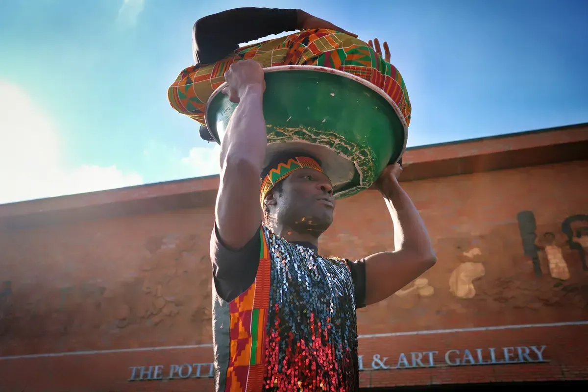 Man carrying a large green bowl on his head, in front of a pottery gallery.