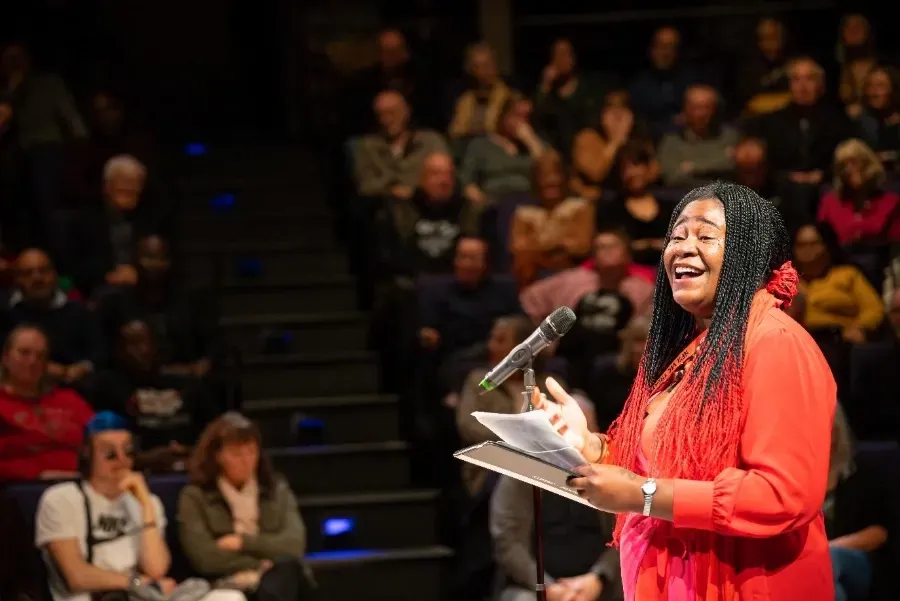Woman in red dress with braids sings at microphone, holding papers, stage setting with audience.