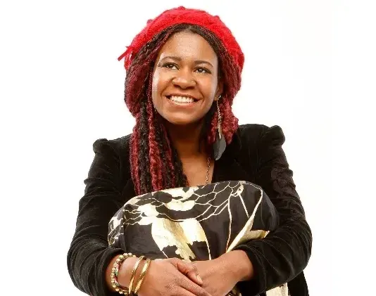 Smiling woman with red-tipped dreadlocks and a red beret holds a black and gold patterned cloth.