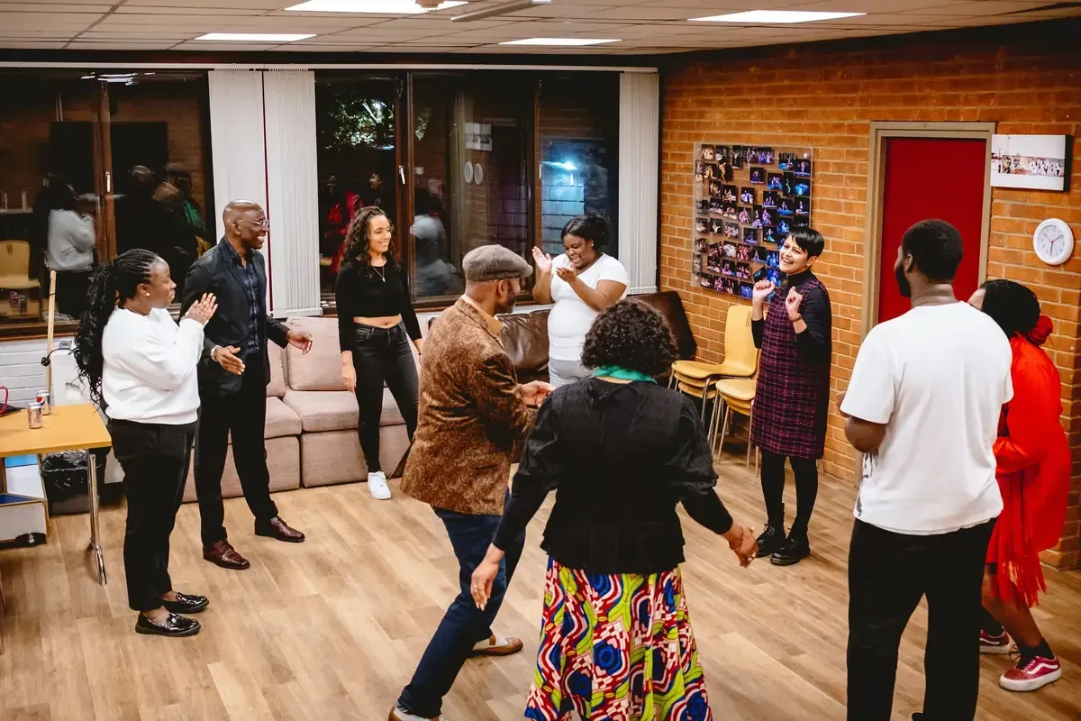 People dancing in a well-lit room with hardwood floors. Some are clapping.