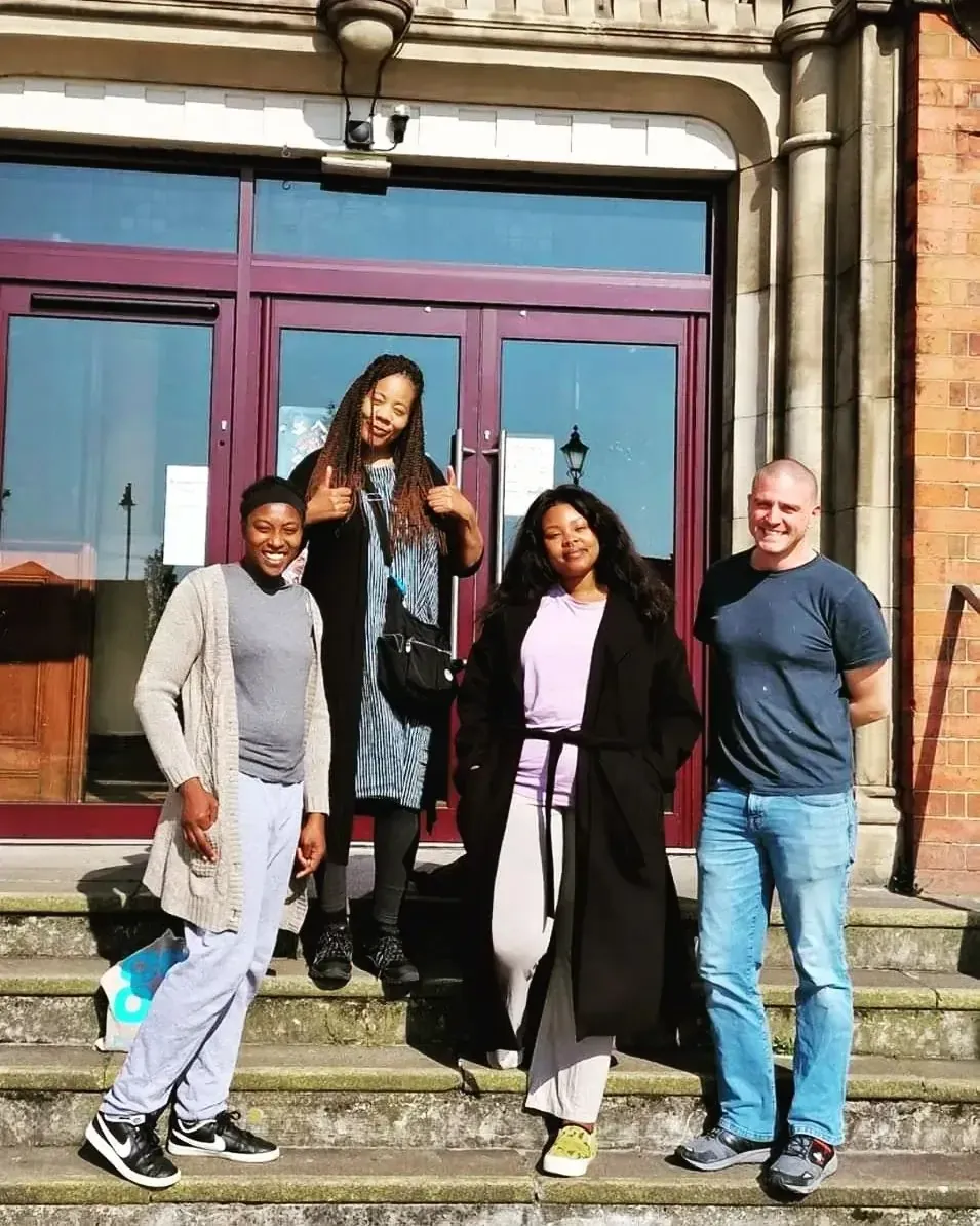 Four people pose on steps of a brick building; two women giving thumbs up. Sunny day.
