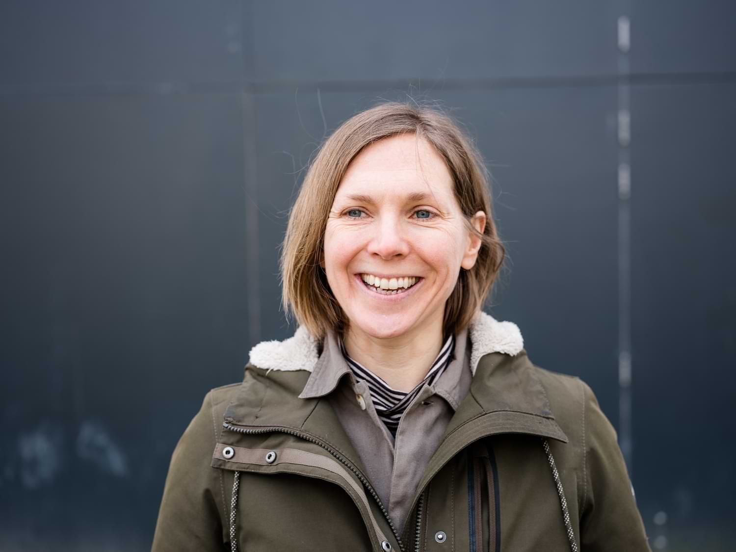 Woman smiles, wearing olive jacket, in front of a dark wall.