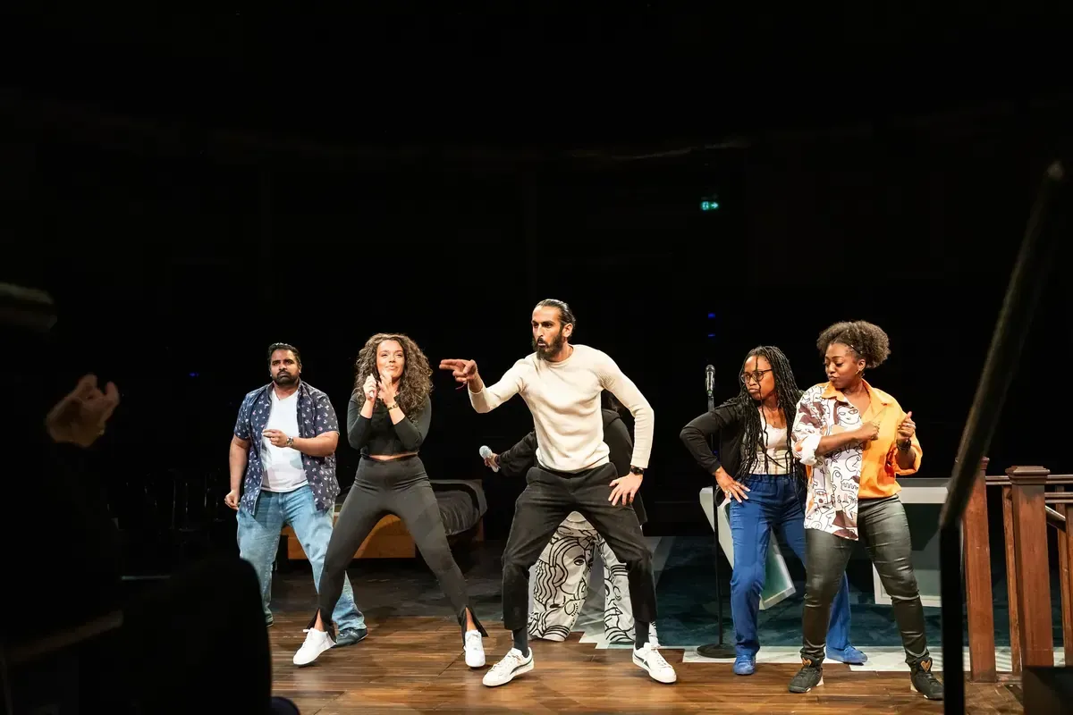 Theater stage with six people dancing. One points a fake gun. Dark background.