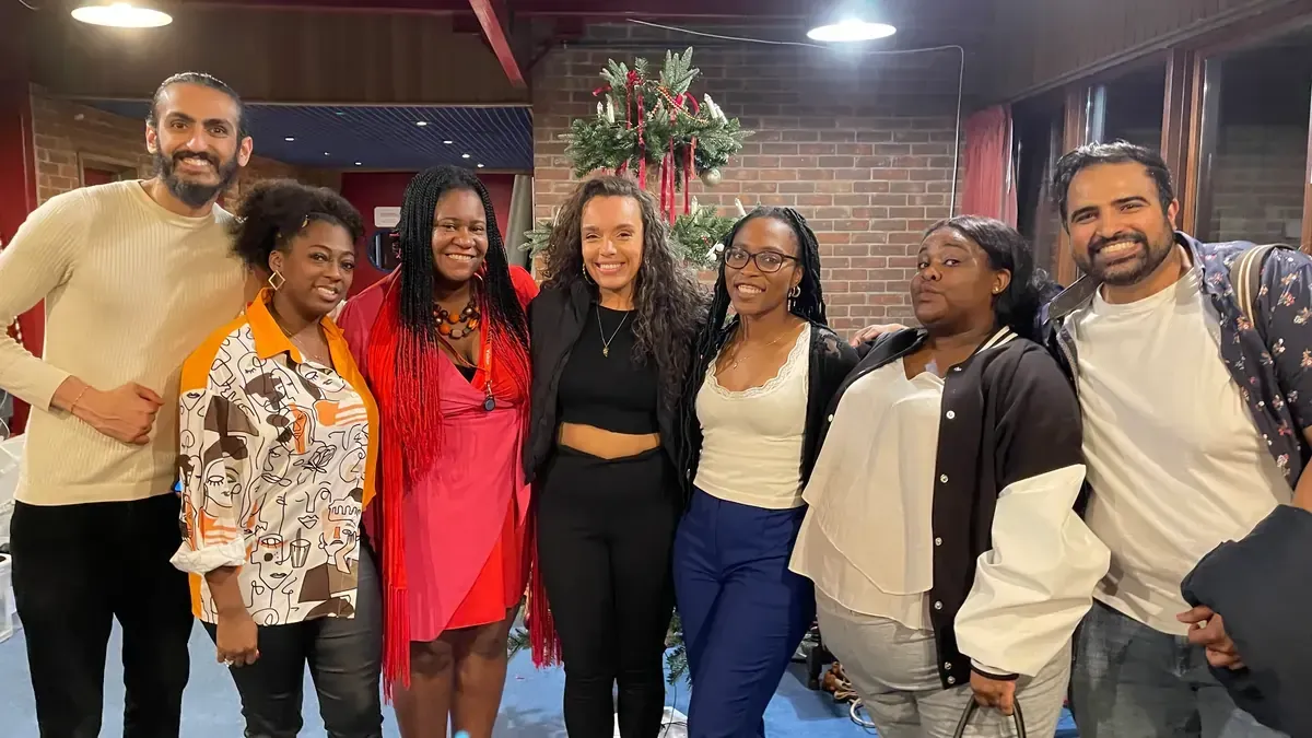 Group of diverse people pose together indoors, smiling. Some have arms around each other; a festive setting.
