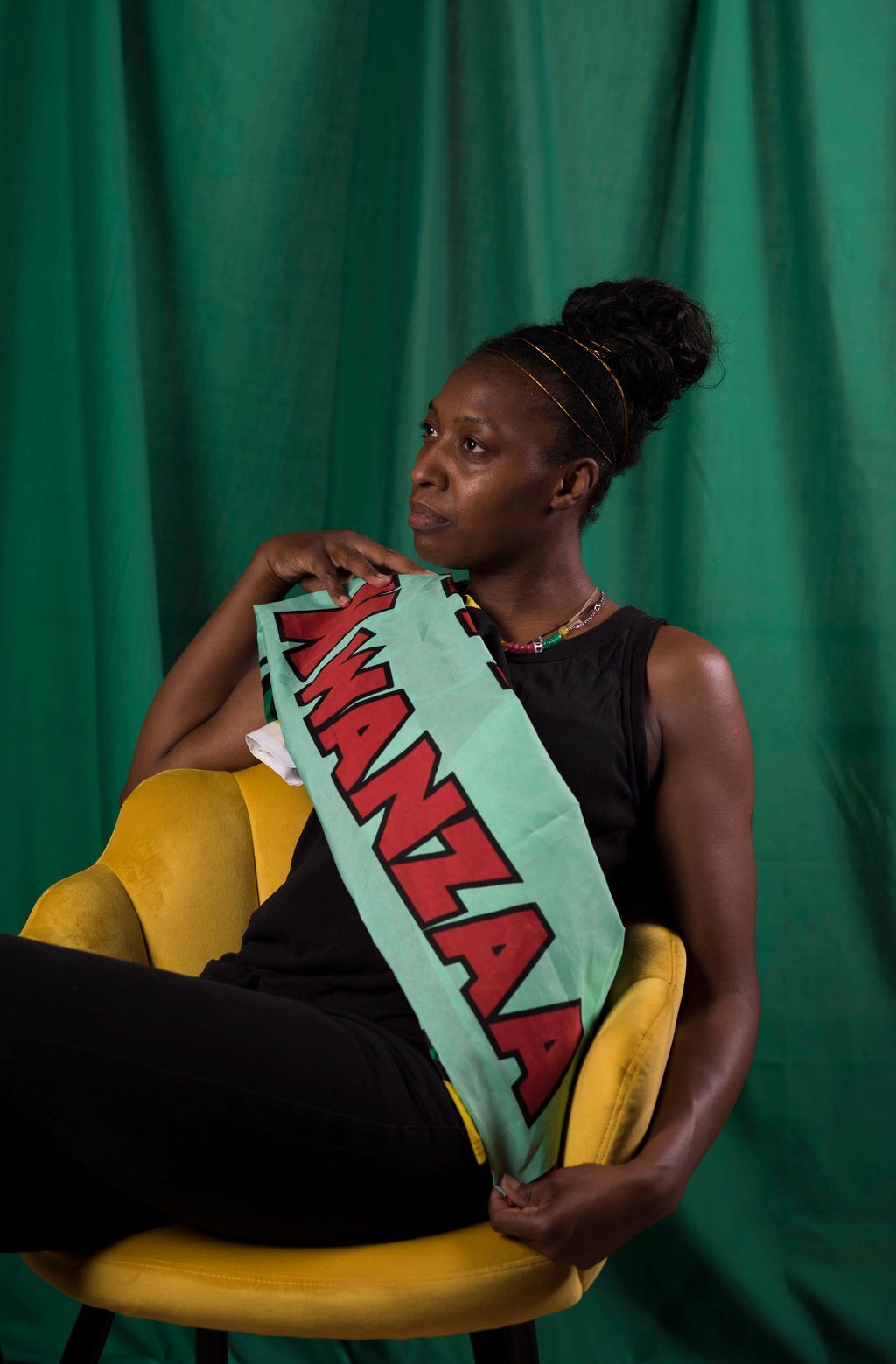 Woman in black outfit seated, holding a sash with "MANZAA" written on it, set against a green backdrop.