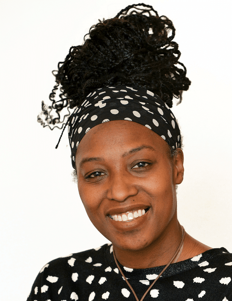 Woman with a black and white polka dot headband and sweater smiles. Dark curly hair is pulled up. White background.