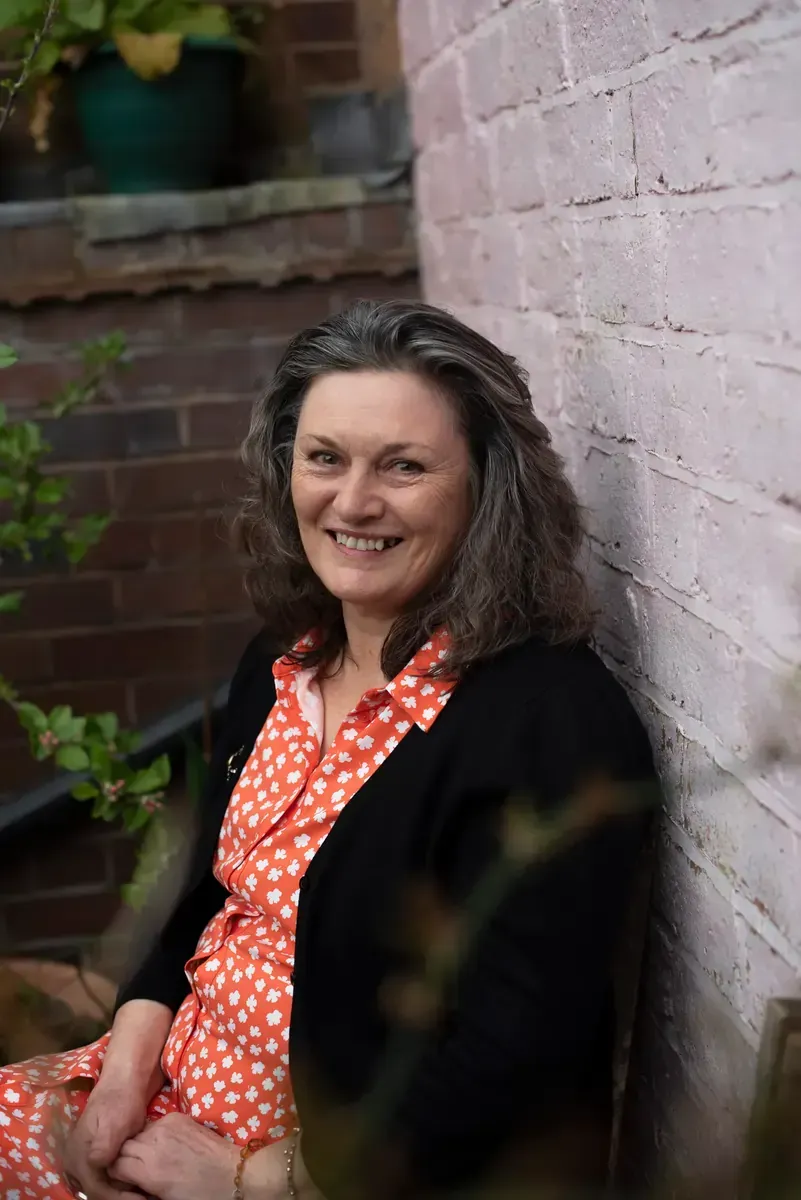 Woman with graying hair smiles, seated outside, leaning against a brick wall. Wearing black sweater and patterned dress.