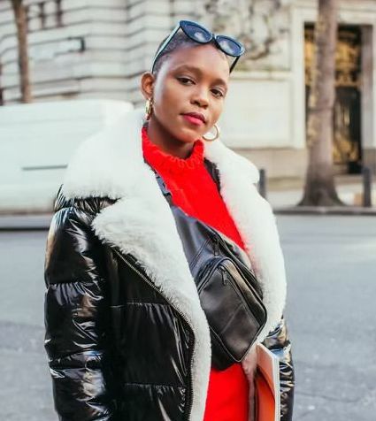 Woman in black puffer jacket with white fur trim, wearing red sweater and sunglasses, standing outside.