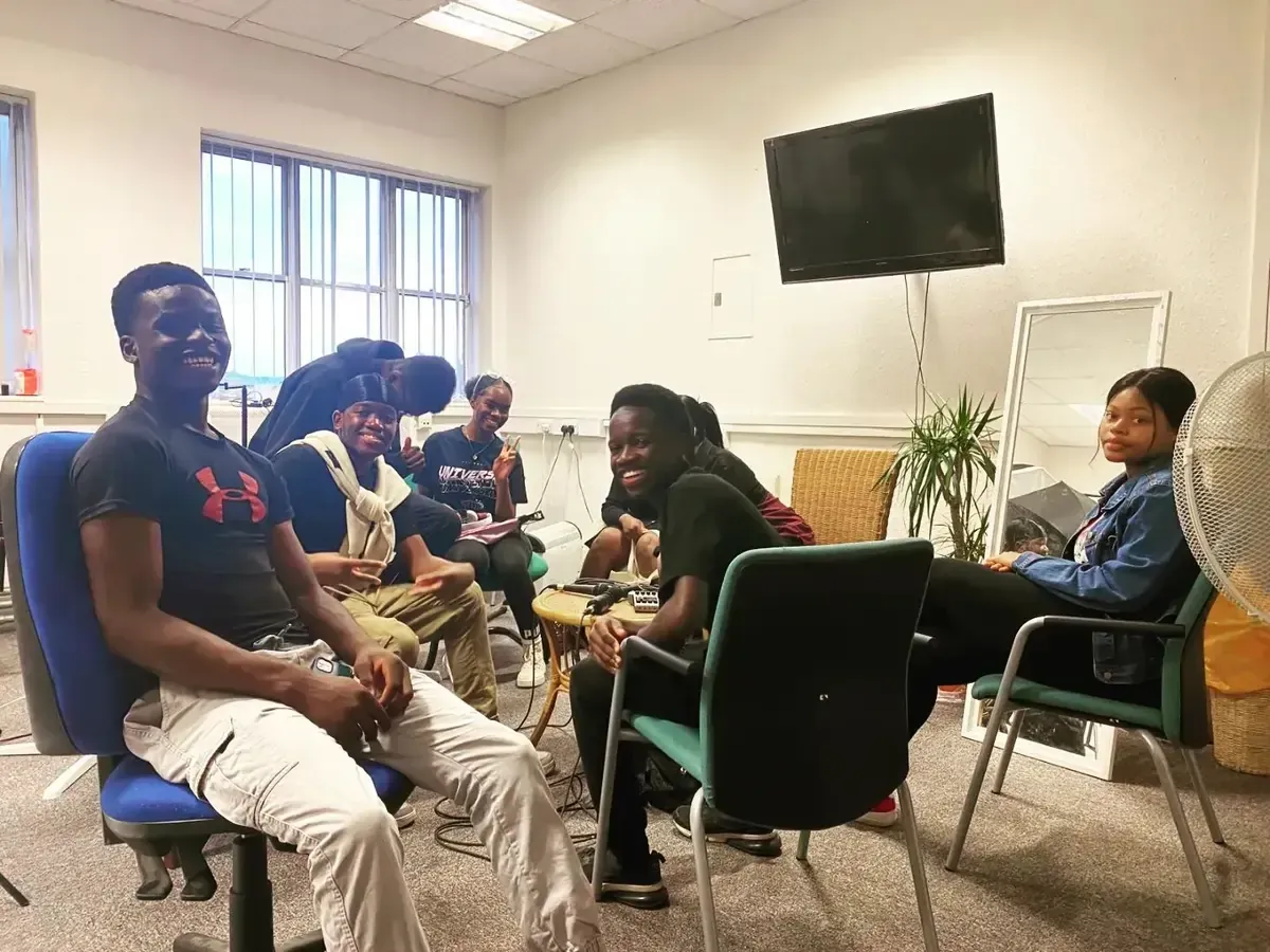 Group of young Black people in a room; some seated, smiling, others interacting. Bright room with a TV, window, and a fan.