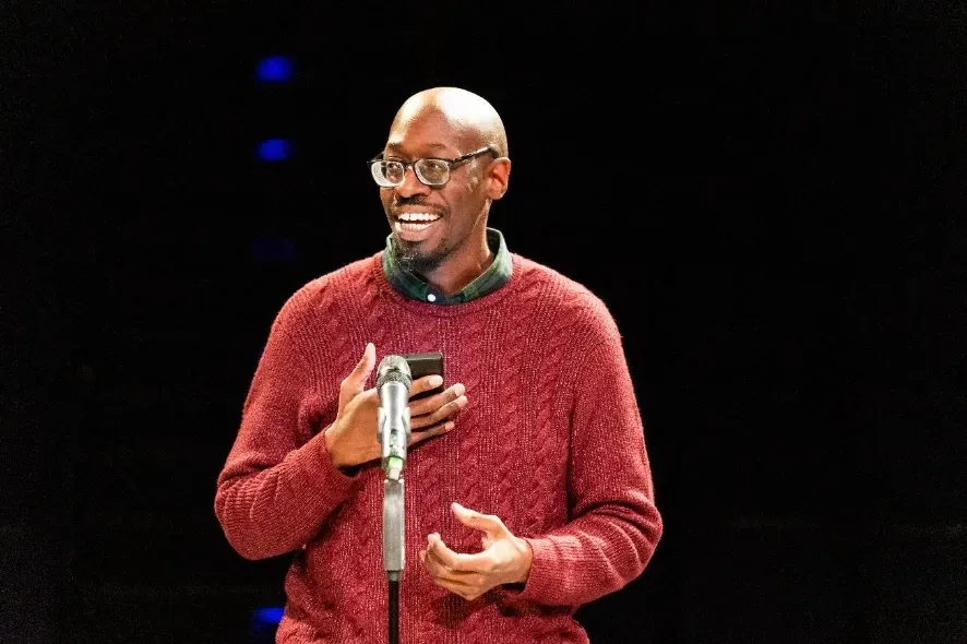 A Black man with glasses smiles, holding a microphone and phone, standing on stage in front of a dark background.