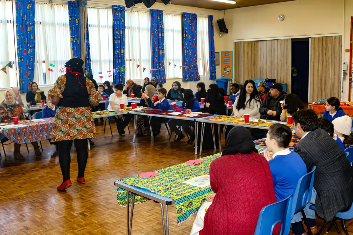 A woman in a colorful dress speaks to a group seated at tables in a decorated hall.