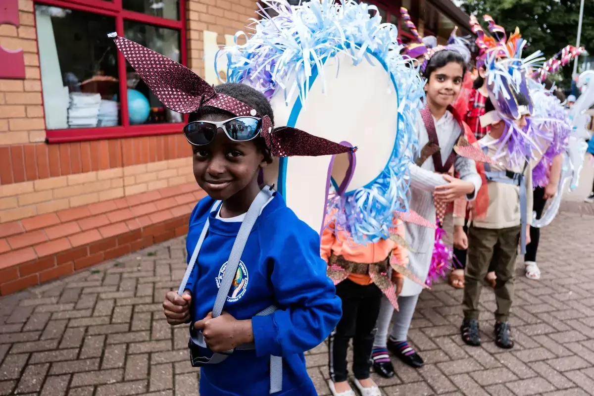 Children in costumes parade, smiling. One wears sunglasses and a blue top. Brick building in background.
