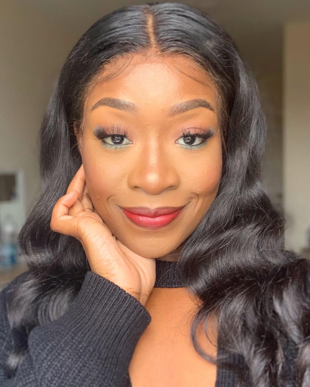 Black woman with long wavy hair, wearing makeup, smiling, and holding her face.