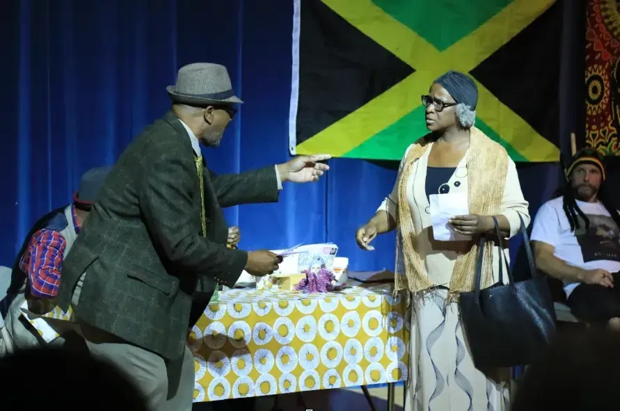 Man handing papers to woman in front of a Jamaican flag, a table, and two other people.