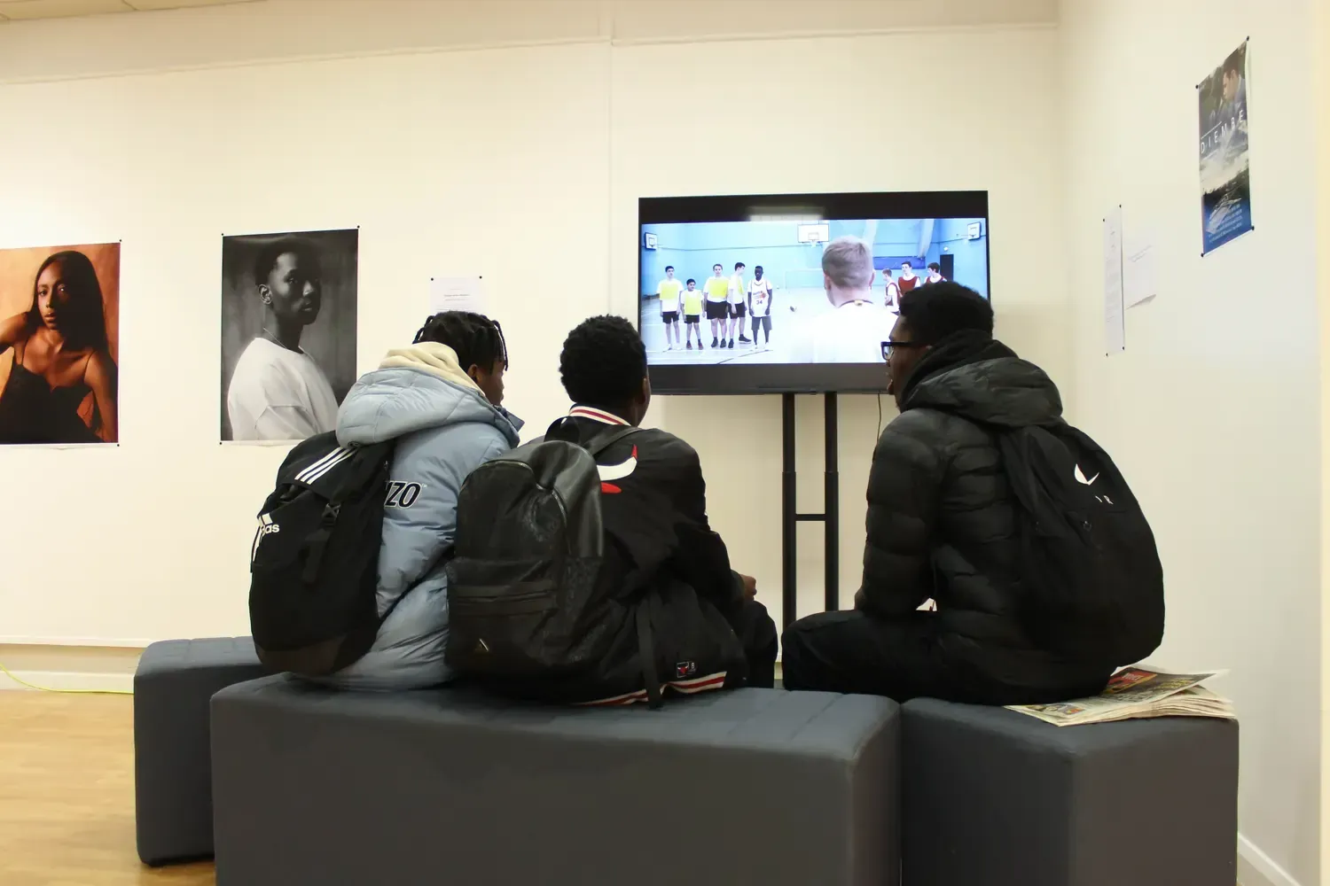 Three people watch TV in a room with photos on the wall. They sit on a gray bench, backs to the camera.