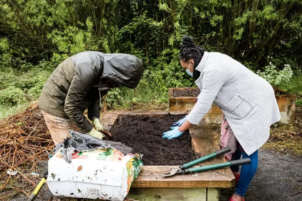 Two people, gloved, working in a raised garden bed, adding soil. Outdoor setting.