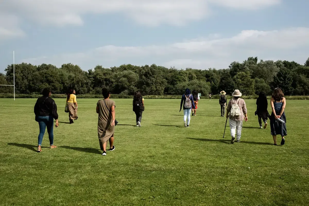People walking on a grassy field under a blue sky. Trees in the background.