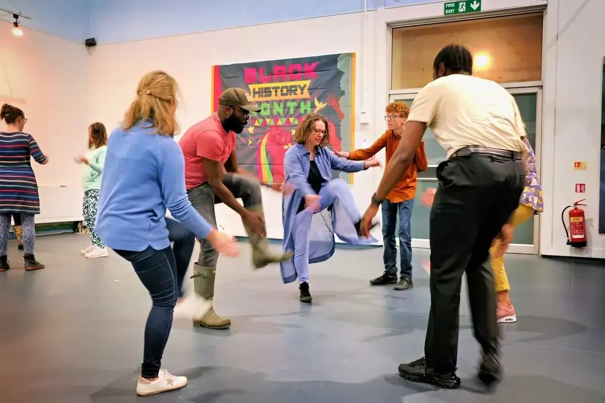 People of various ages dancing in a room; Black History Month poster visible in the background.