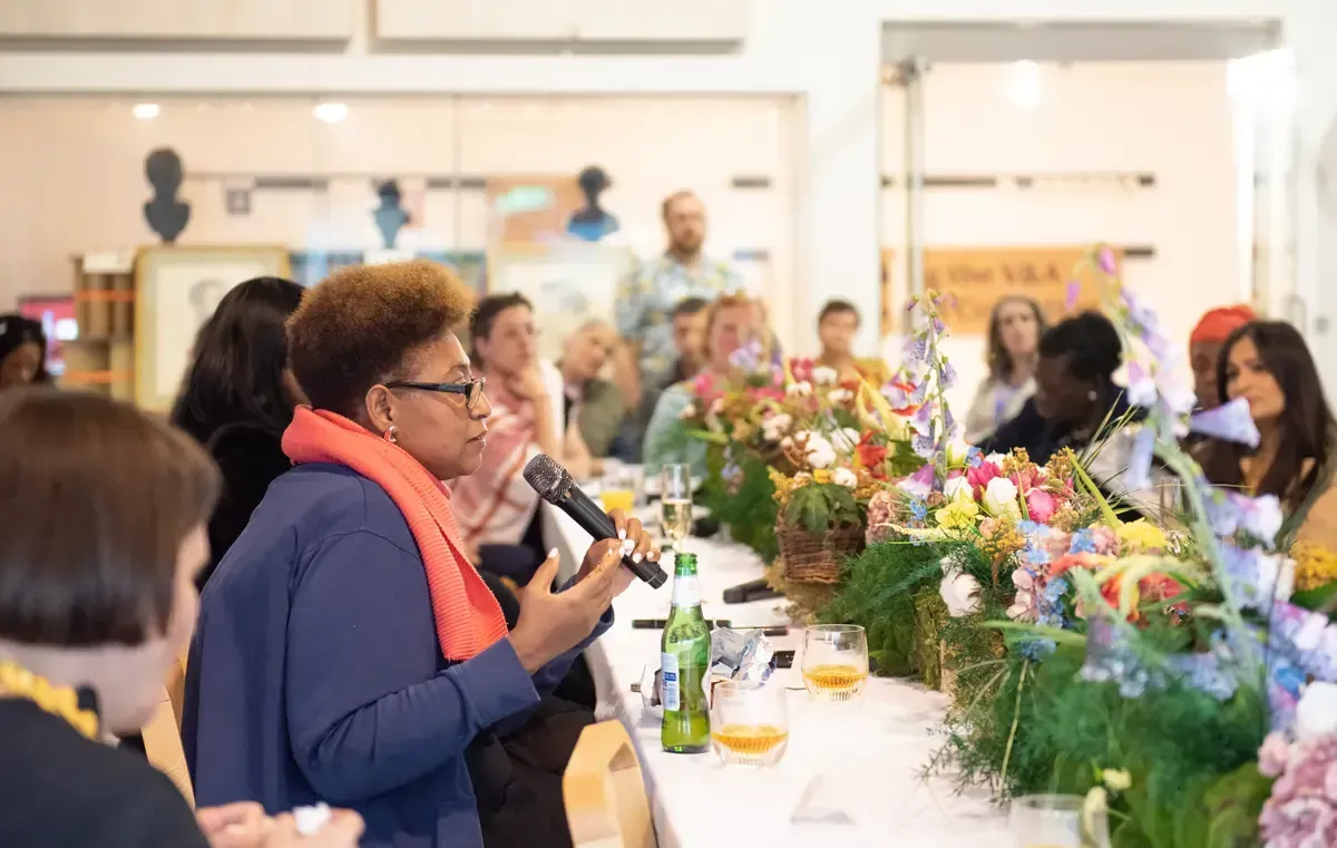 Woman with microphone speaks at a table with floral arrangements. People listen at a long table.
