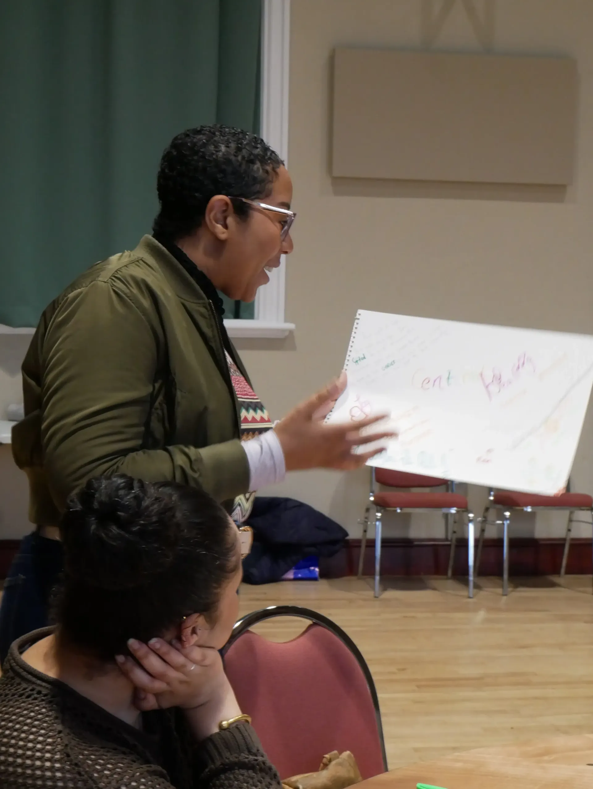 Woman in green jacket gestures, holding a whiteboard during a presentation. Another woman listens.