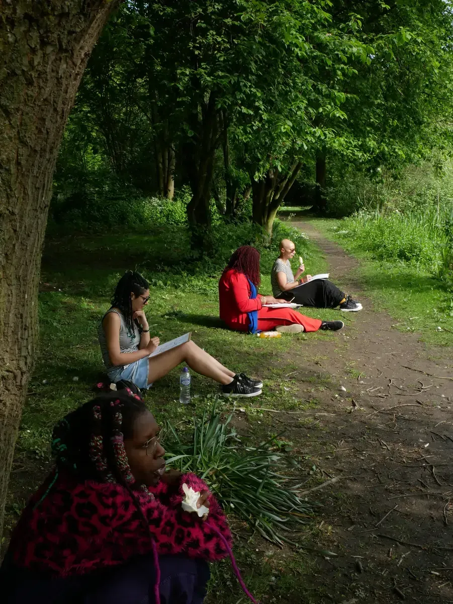 Four people sketching outdoors in a park: a path, trees, sunlight, and grass.