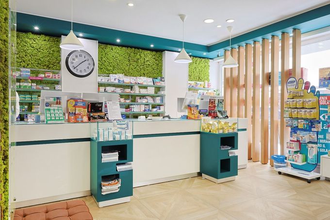 Luminoso interno di farmacia con scaffali pieni di prodotti, finiture color verde acqua e una parete ricoperta di muschio verde con panca.
