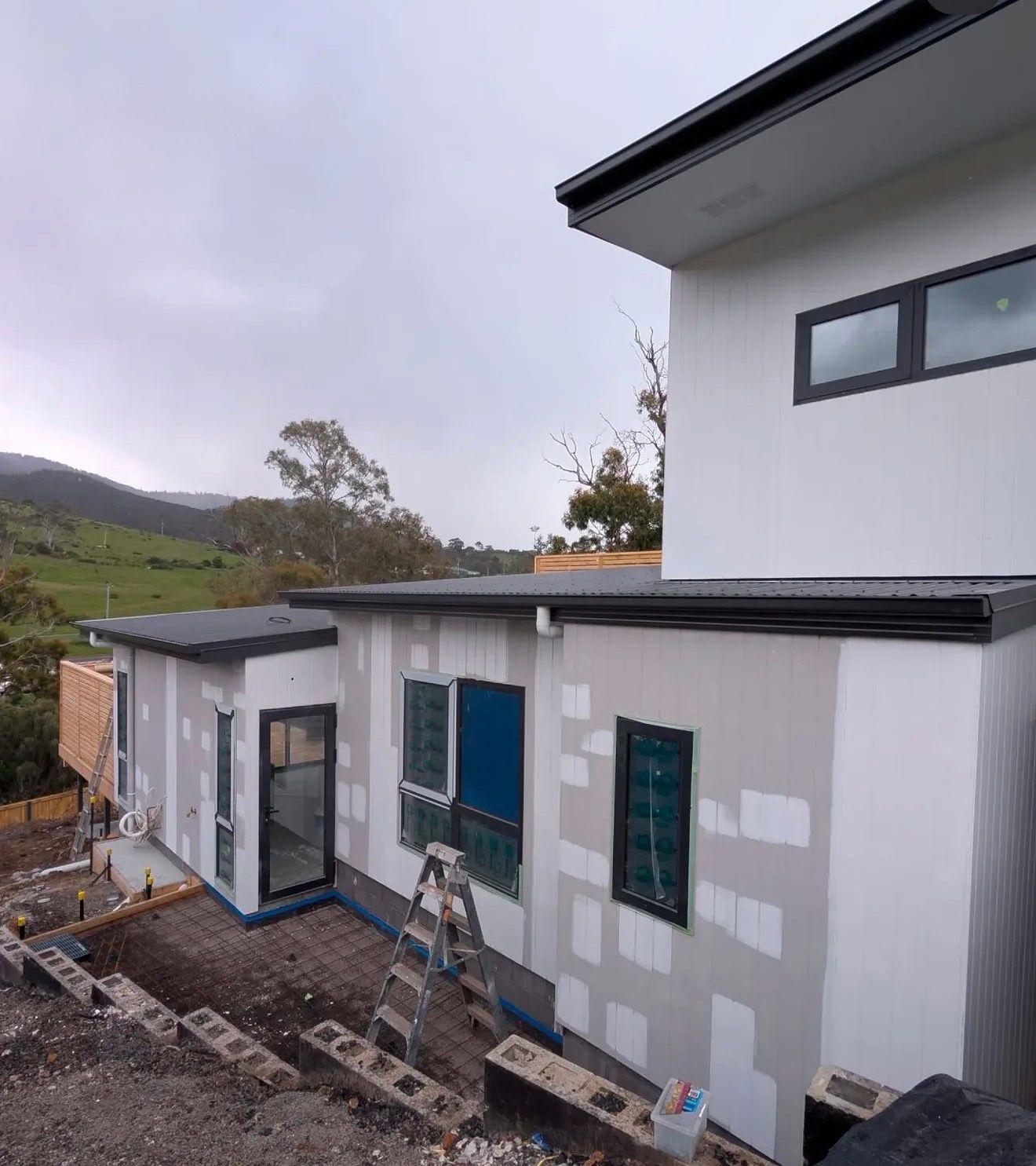 A modern, two-story house under construction with white exterior cladding, black window frames, and a metal roof.