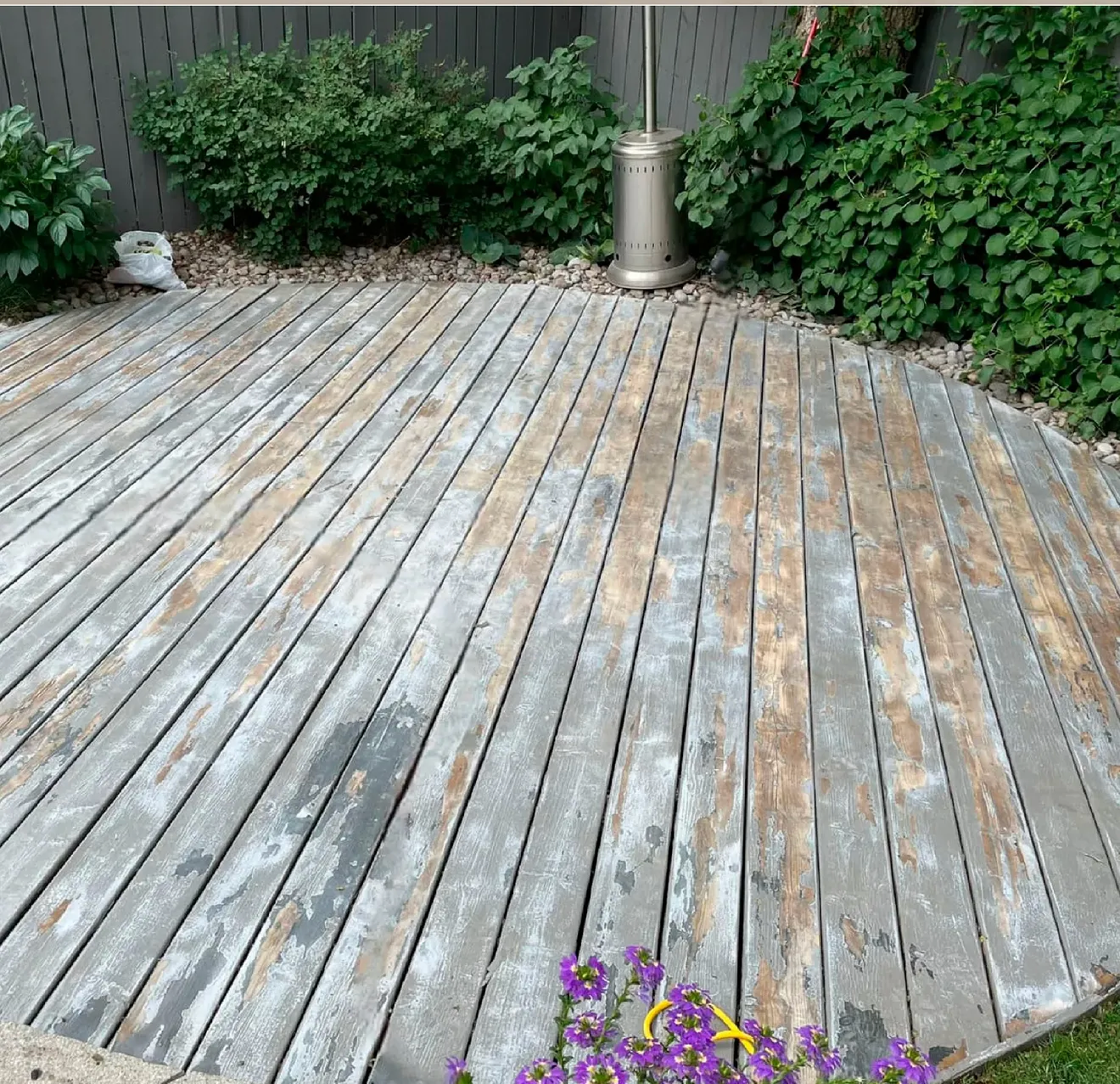A circular wooden deck in a backyard featuring weathered grey planks, surrounded by green shrubs and a metal heater.