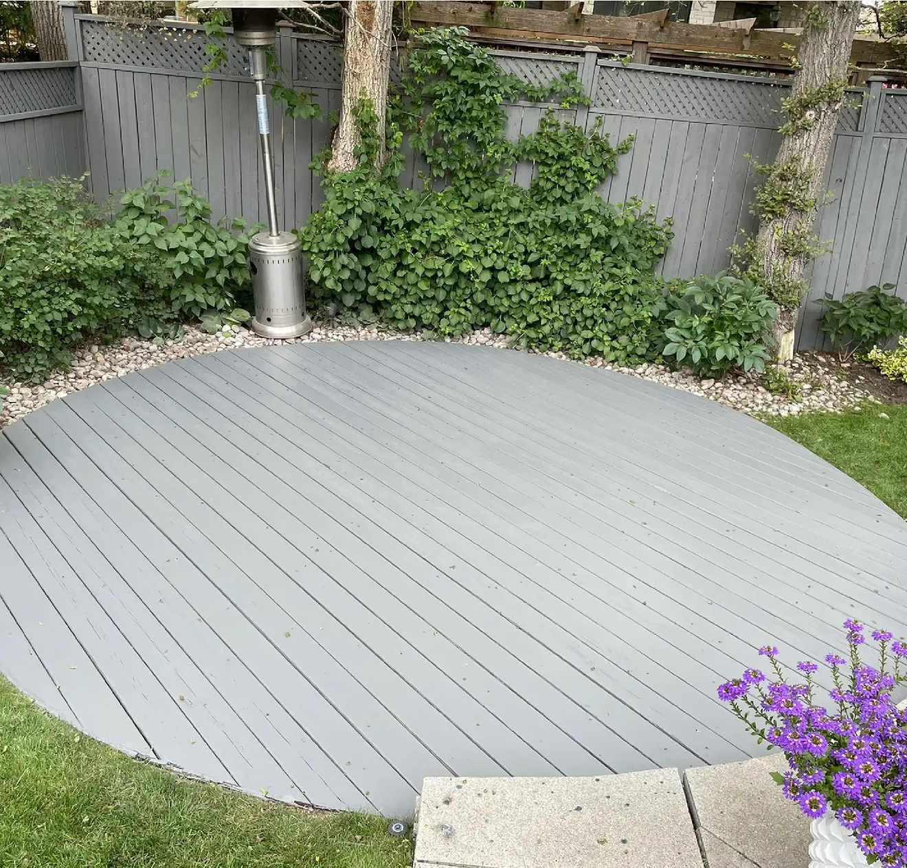 A circular grey wooden deck sits in a backyard next to a metal patio heater, green bushes, and a lattice fence.