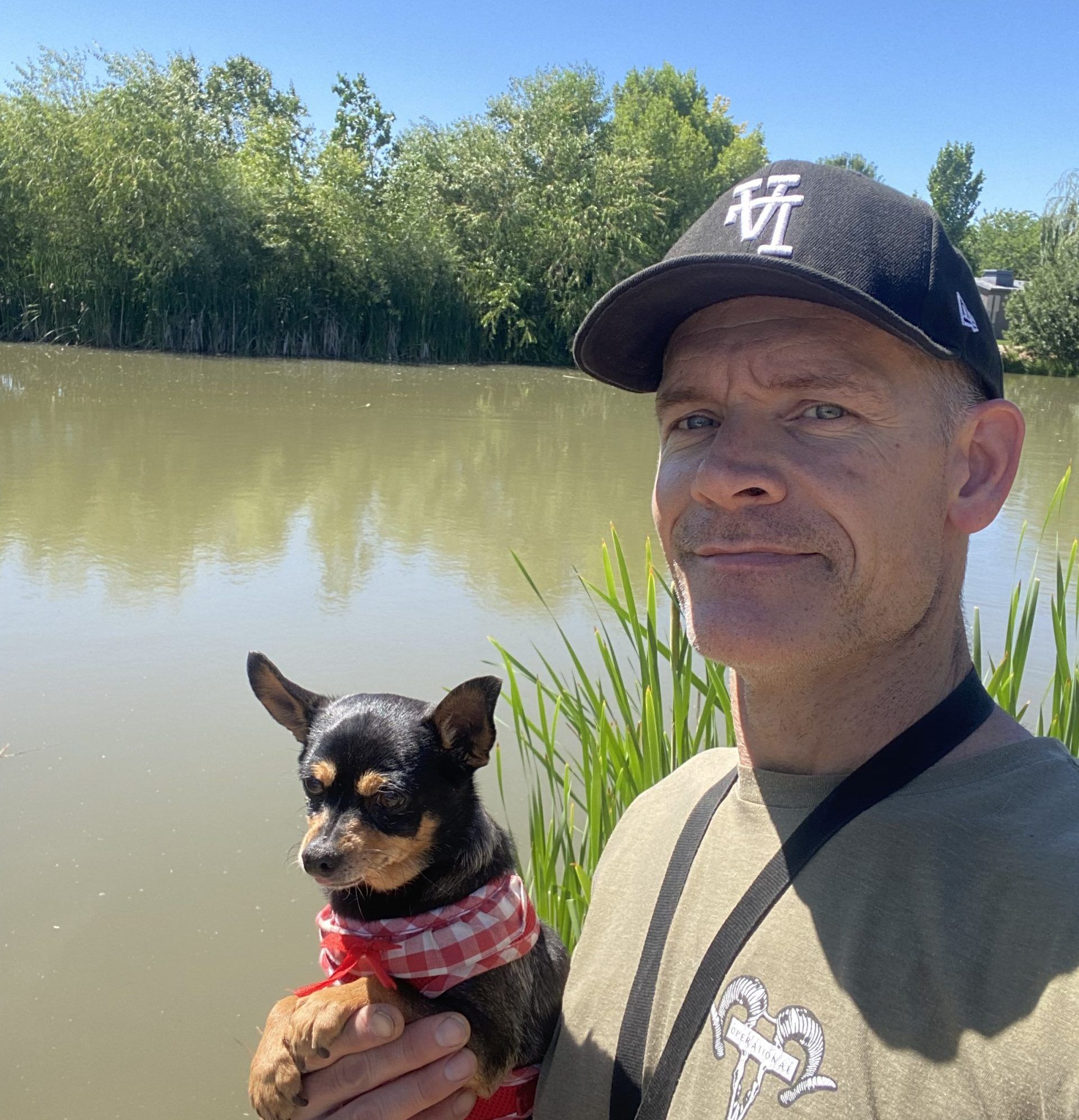 Man holding a small dog by a calm river, with bamboo and blue sky in the background.