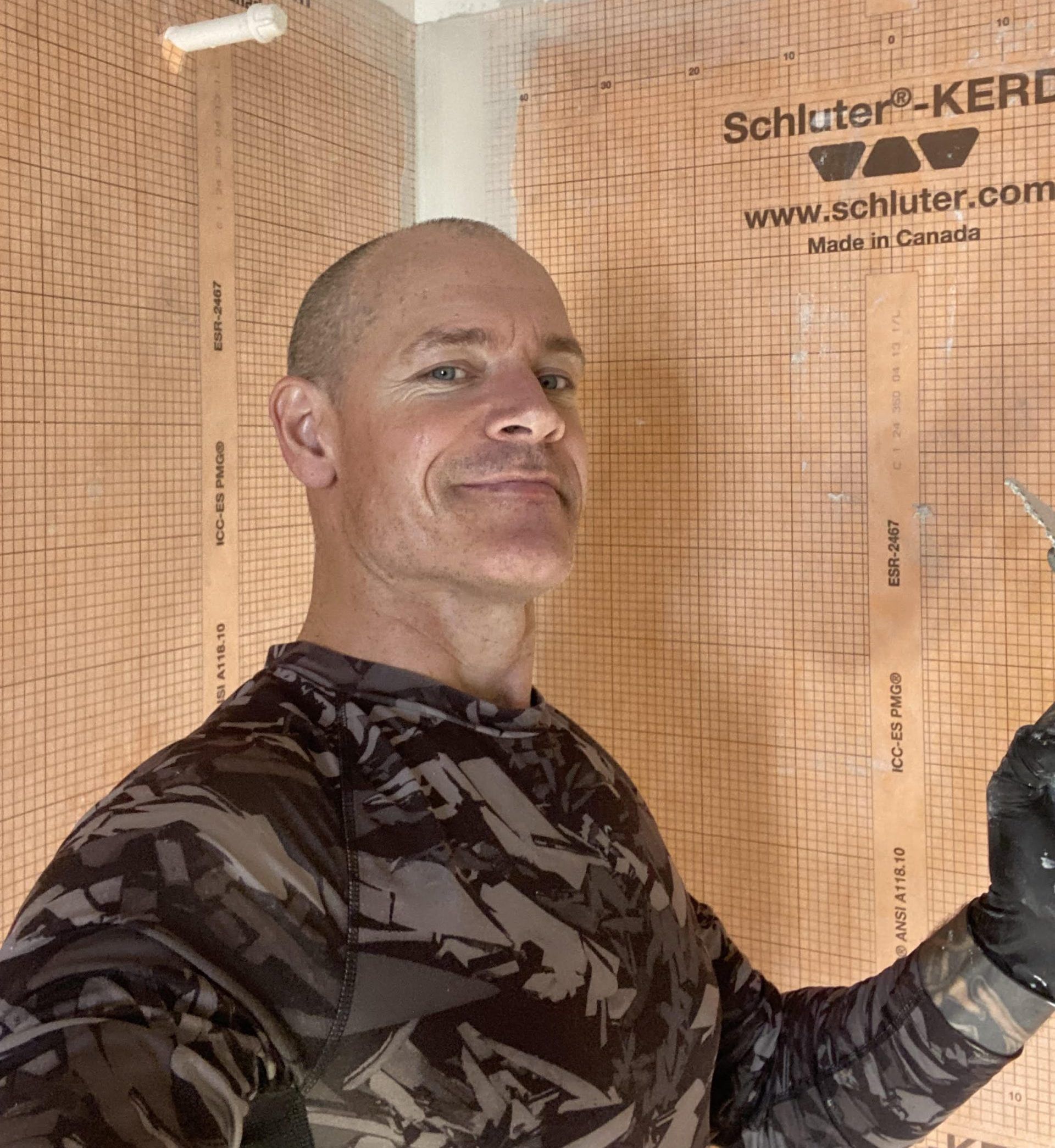 Man in camouflage shirt smiling inside a tiled construction area, holding a tool or cable.