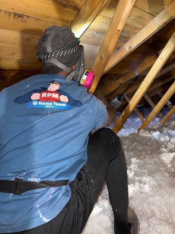 Person in attic, wearing respirator and RPM Home Team shirt, working among insulation.