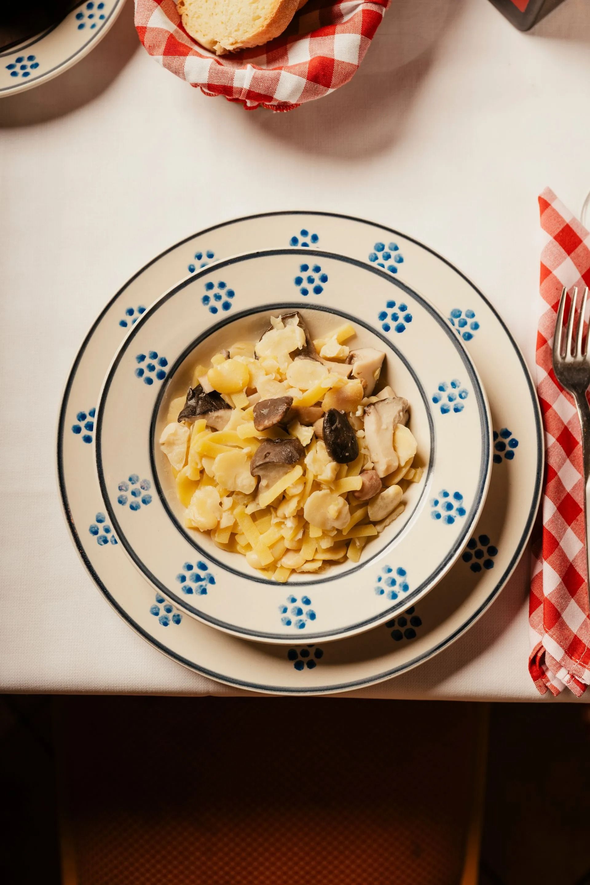 Un piatto di pasta cremosa ai funghi su un piatto blu decorato con fiori, adagiato su una tovaglia a quadri, con del pane a portata di mano.