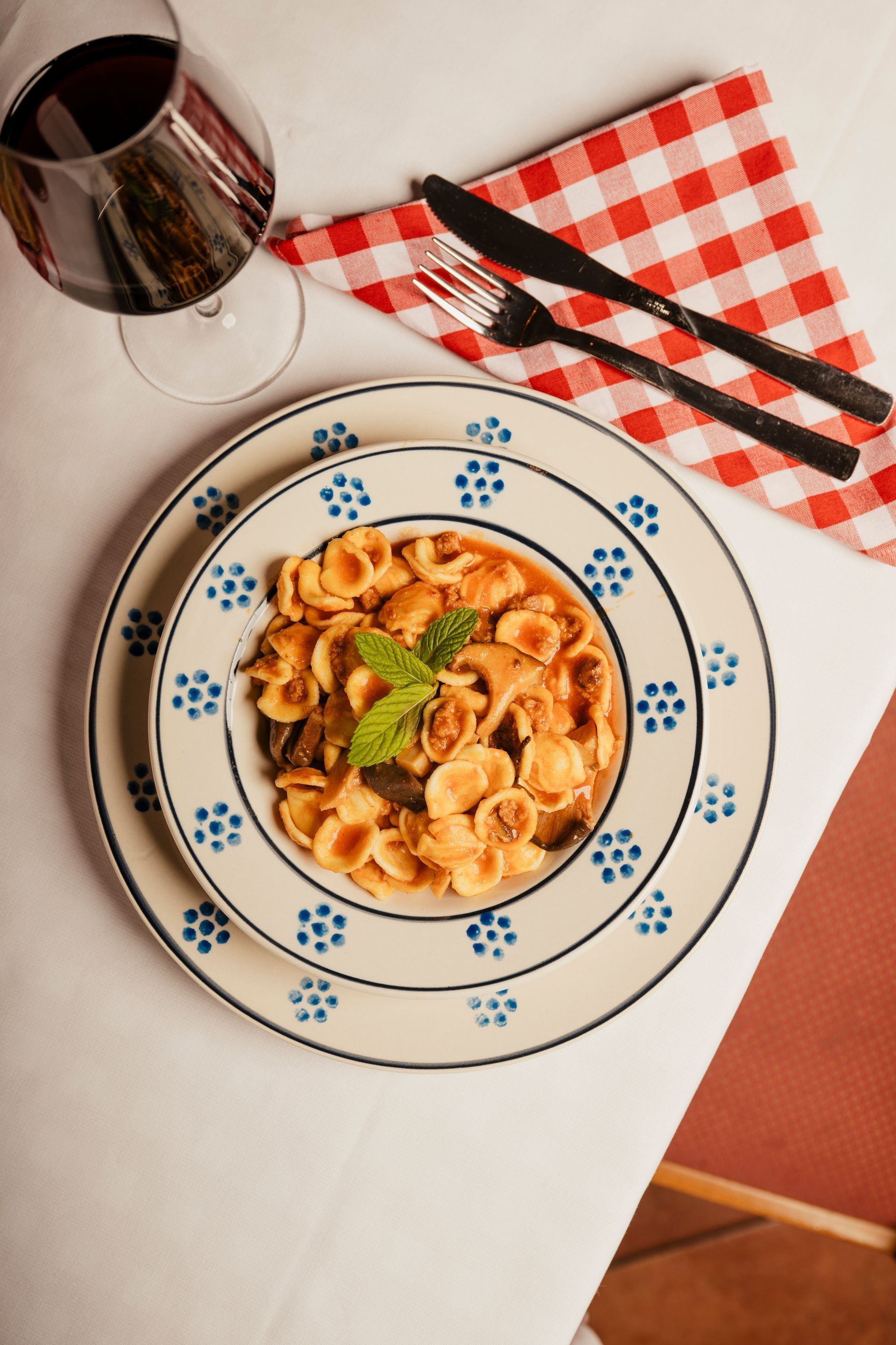 Un piatto di pasta su un piatto con il bordo blu, accanto a un bicchiere di vino rosso e un tovagliolo a quadretti su un tavolo.