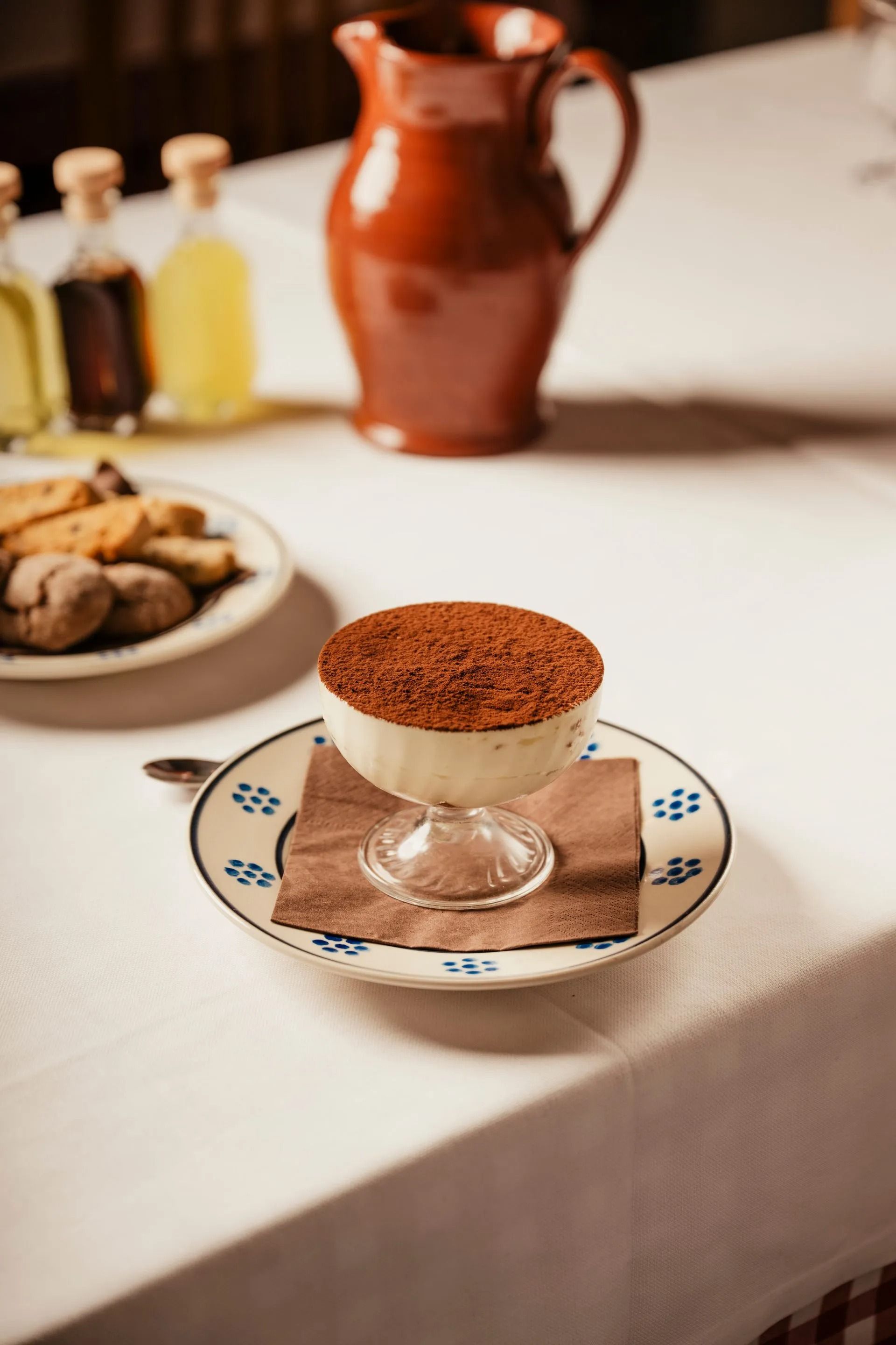 Dessert in un bicchiere su un piatto decorato, con una caffettiera e biscotti su un tavolo.