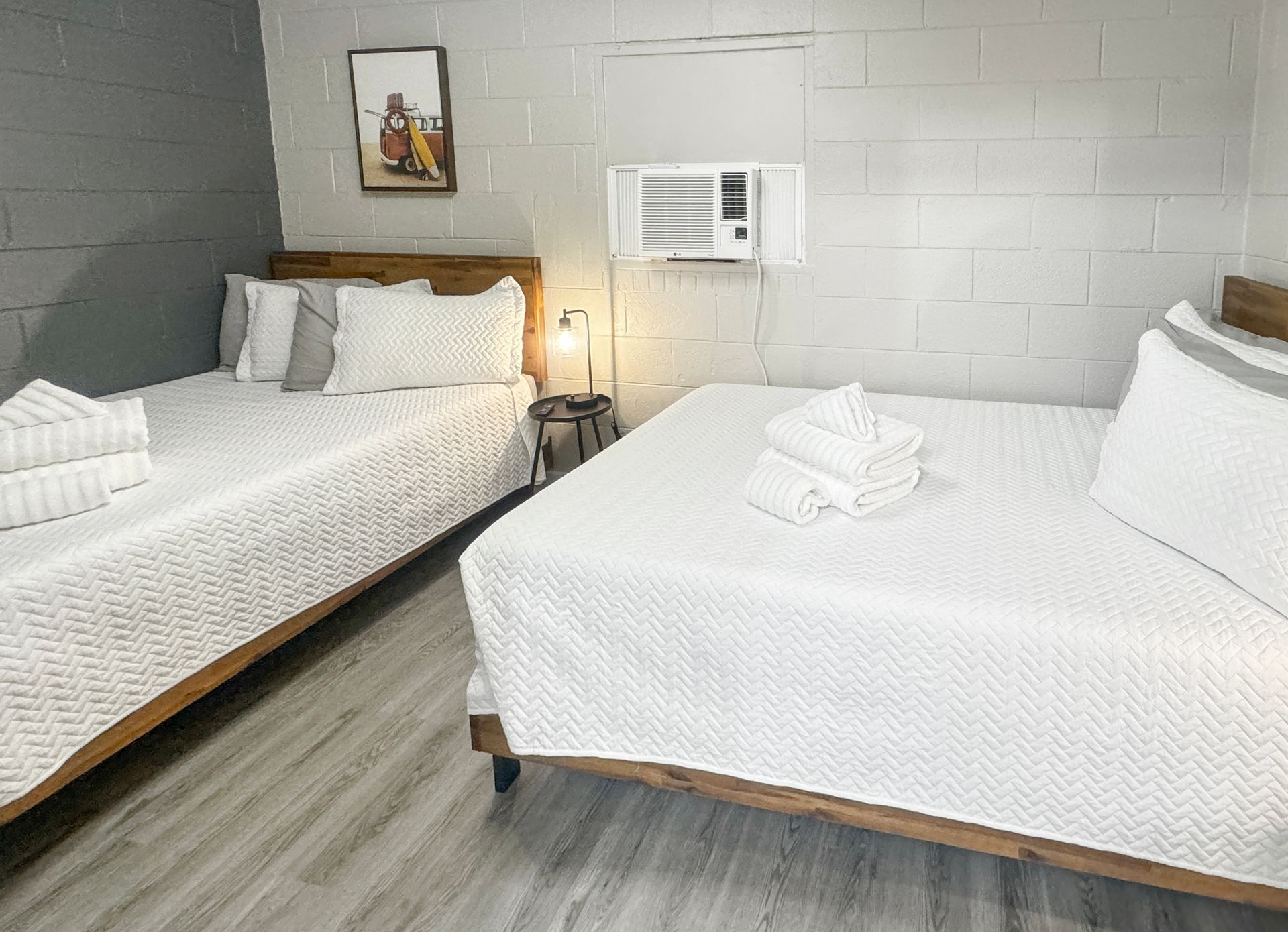 Two beds in a motel room with white bedding, a window air conditioner, and a framed picture.